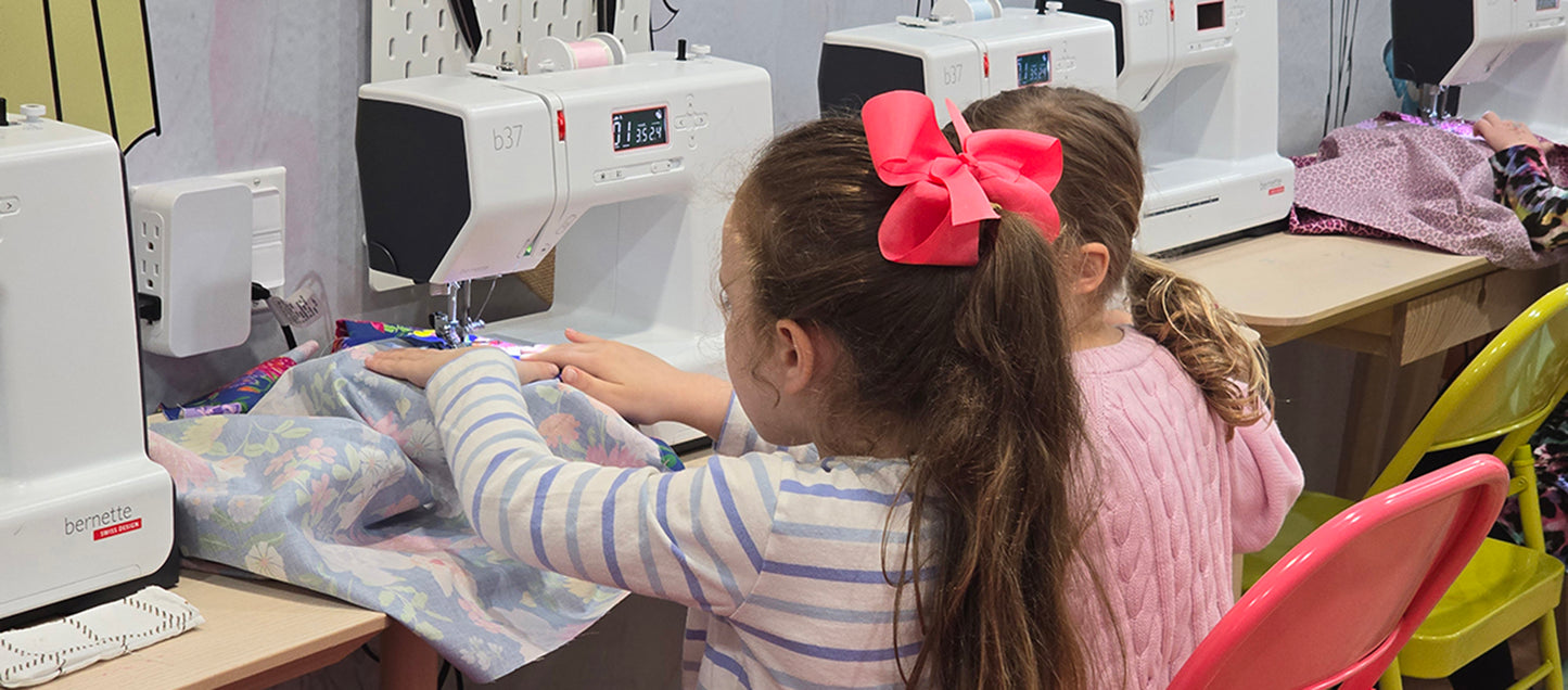 Two young girls working on a sewing project with multiple sewing machines in the background.
