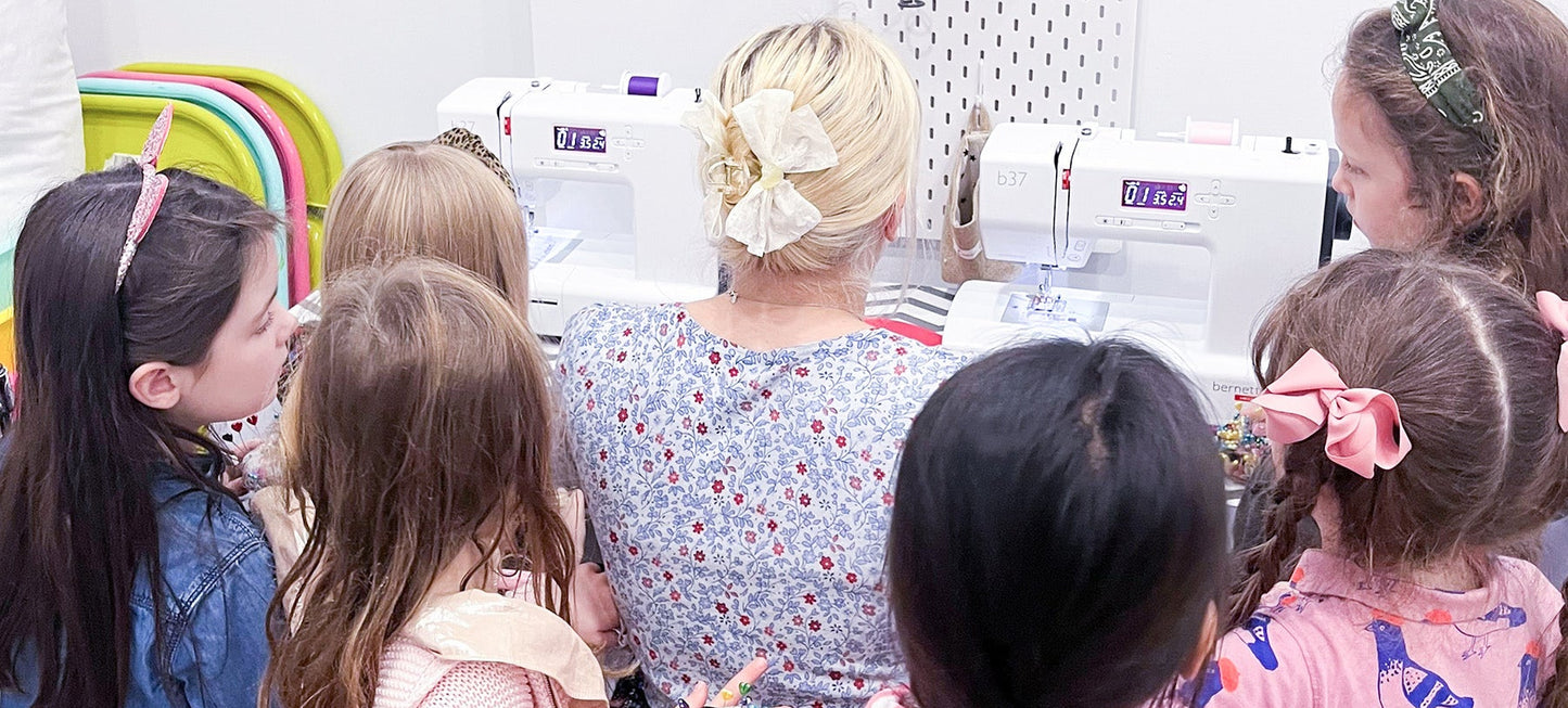 Group of children and a teacher at a sewing machine in a classroom setting.